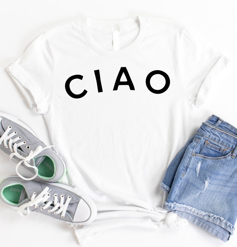 May include: White t-shirt with the word "CIAO" printed in black on the front. The shirt is laid flat on a white surface with a pair of gray sneakers and blue denim shorts.