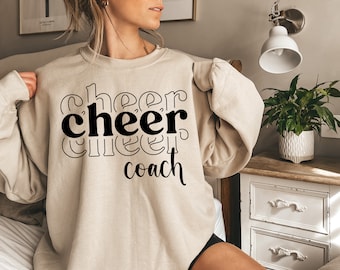Peut inclure: Un sweat-shirt beige avec le texte "cheer coach" imprimé en noir.