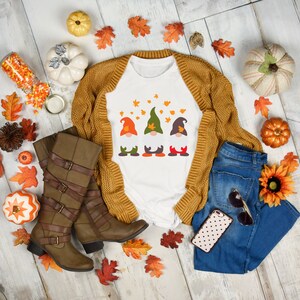 May include: White t-shirt with a graphic of three gnomes wearing fall colors and surrounded by autumn leaves. The shirt is paired with a brown cardigan, blue jeans, brown boots, and fall-themed decor.