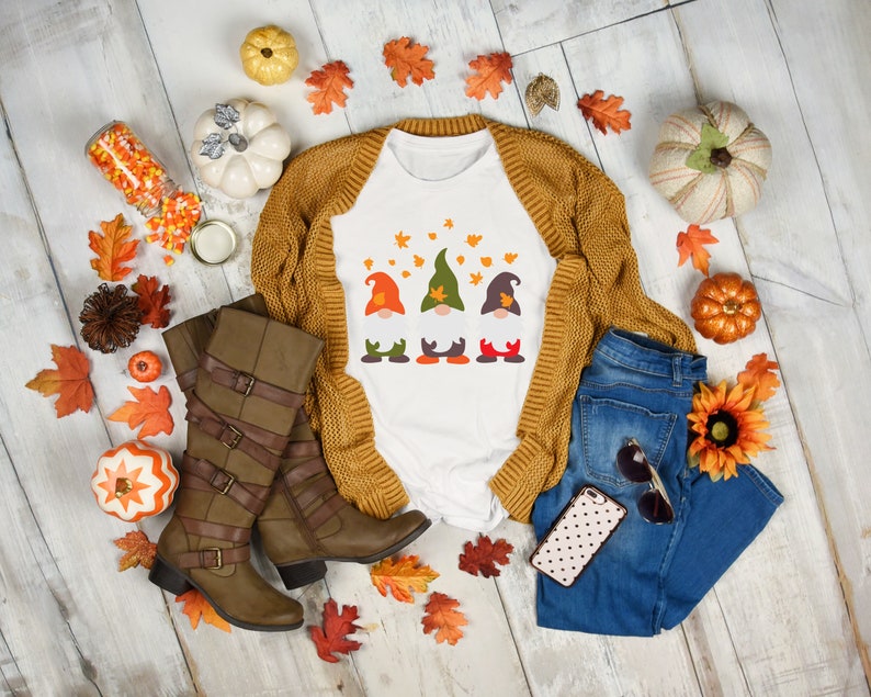 May include: White t-shirt with a graphic of three gnomes wearing fall colors and surrounded by autumn leaves. The shirt is paired with a brown cardigan, blue jeans, brown boots, and fall-themed decor.