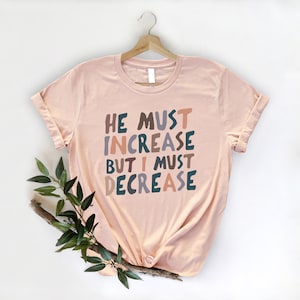May include: A peach-coloured t-shirt with the text "He must increase but I must decrease" in colourful, stylised lettering.