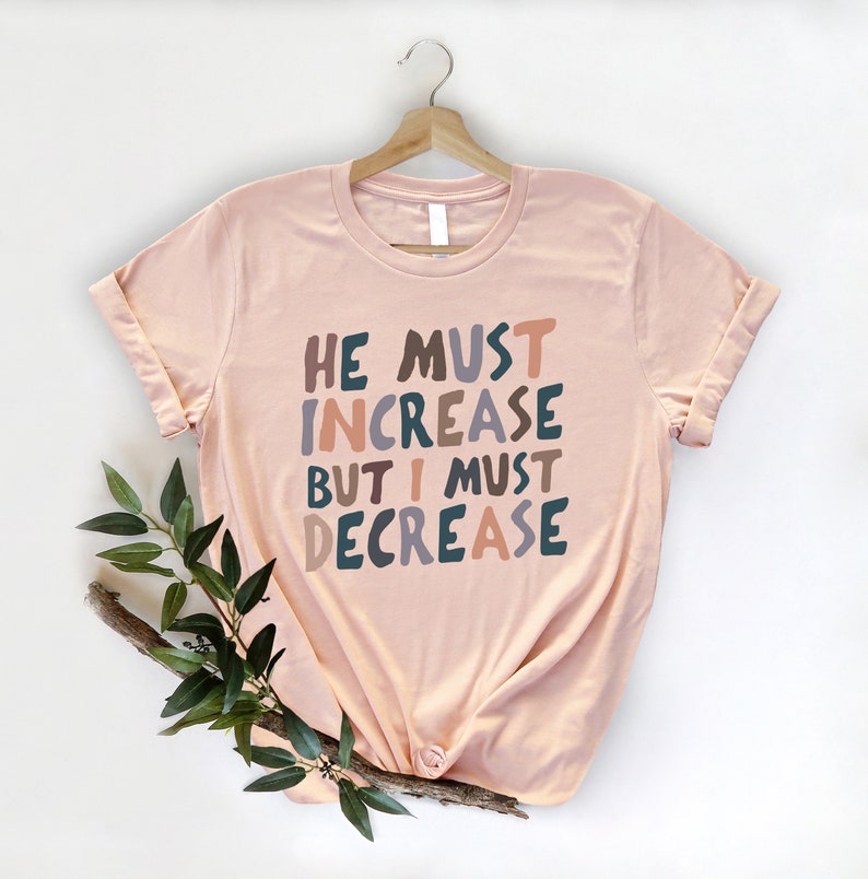 K&ouml;nnte beinhalten: Ein pfirsichfarbenes T-Shirt mit dem Text "He must increase but I must decrease" in bunten, stilisierten Buchstaben.