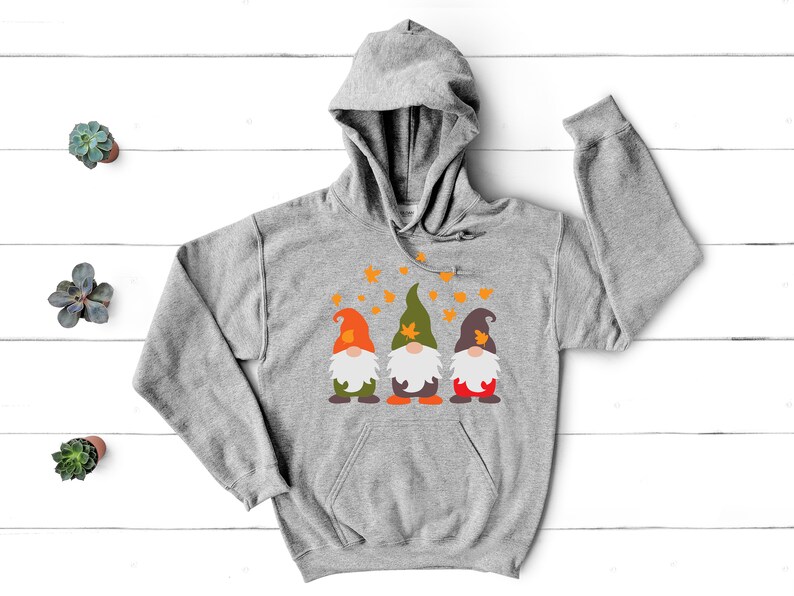 May include: A gray hooded sweatshirt with a design of three gnomes wearing hats with leaves on them. The gnomes are standing in front of a white background with orange leaves falling around them.