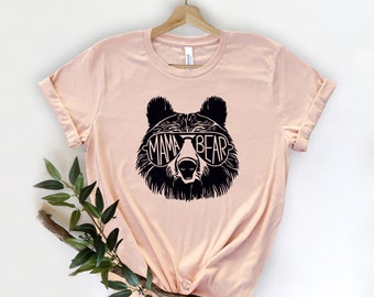 May include: A peach-colored t-shirt with a black graphic of a bear wearing sunglasses. The sunglasses have the words "Mama Bear" written on them.
