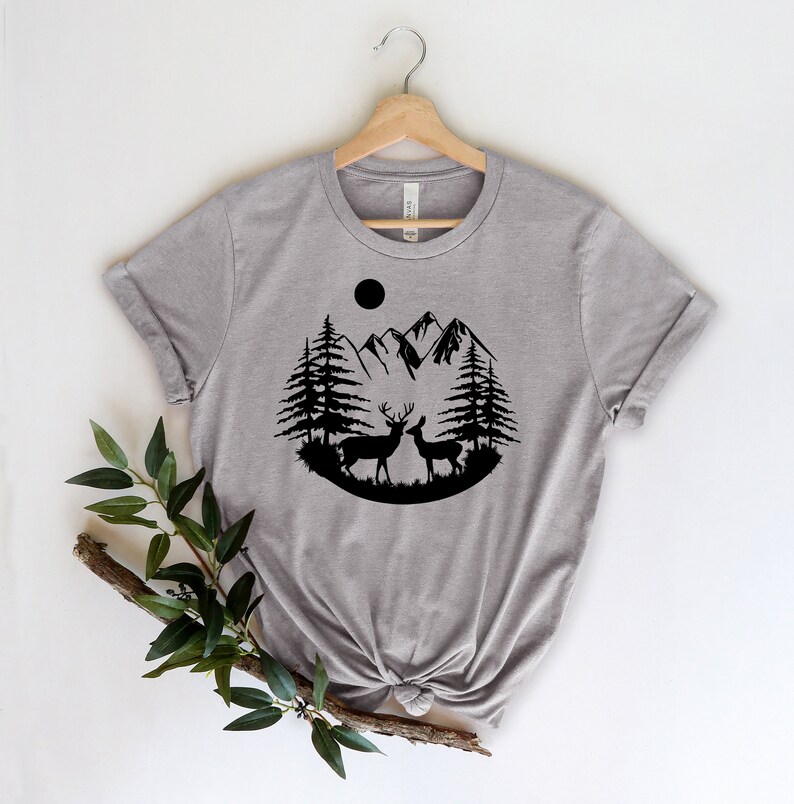 Puede incluir: Una camiseta gris jaspeado con un dise&ntilde;o de silueta negra de dos ciervos, pinos, monta&ntilde;as y un sol. El dise&ntilde;o est&aacute; centrado en la camiseta, con las mangas enrolladas. La camiseta est&aacute; colgada en una percha de madera.