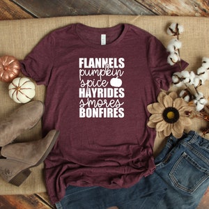 May include: Burgundy t-shirt with white text that reads "Flannels, pumpkin spice, hayrides, smores, bonfires". The shirt is lying on a hessian surface with a pair of brown ankle boots, a pair of blue jeans, and autumnal decorations.