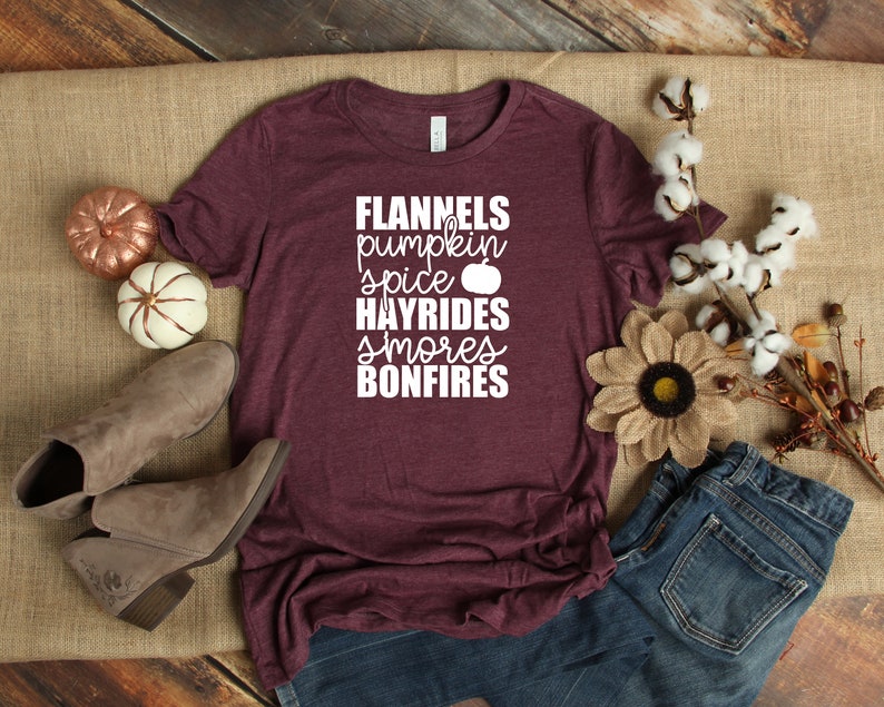 May include: Burgundy t-shirt with white text that reads "Flannels, pumpkin spice, hayrides, smores, bonfires". The shirt is lying on a hessian surface with a pair of brown ankle boots, a pair of blue jeans, and autumnal decorations.