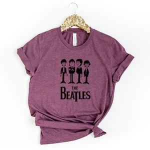 Puede incluir: Una camiseta de color granate con un gr&aacute;fico negro de los Beatles. El gr&aacute;fico presenta una imagen de dibujos animados de los miembros de la banda con trajes, con el texto "THE BEATLES" debajo.