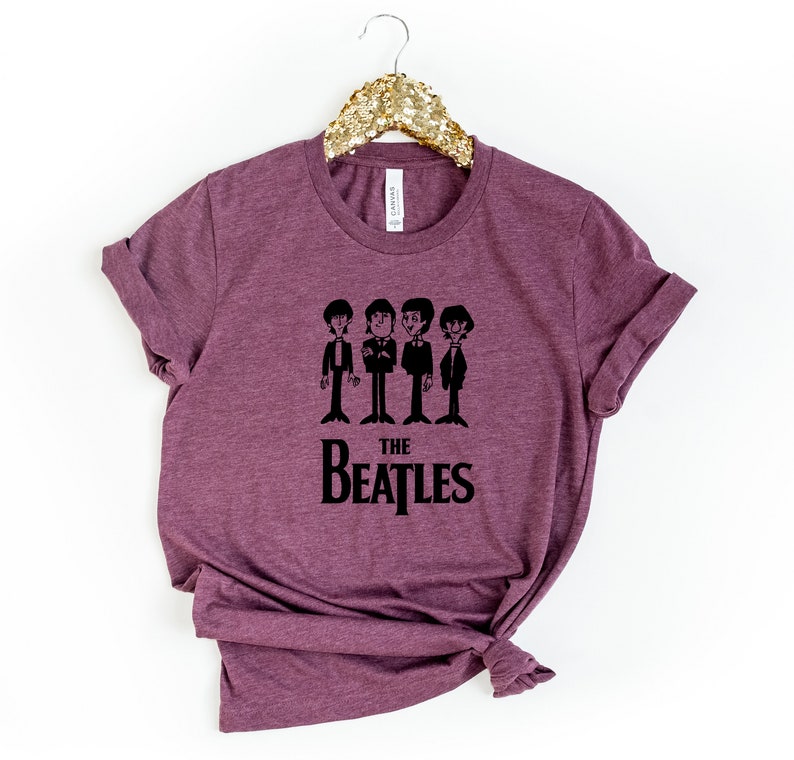 Puede incluir: Una camiseta de color granate con un gr&aacute;fico negro de los Beatles. El gr&aacute;fico presenta una imagen de dibujos animados de los miembros de la banda con trajes, con el texto "THE BEATLES" debajo.