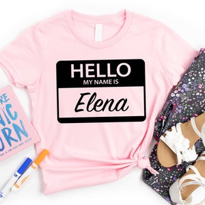May include: Pink t-shirt with a black name tag that says "Hello, My Name Is Elena".