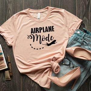 Puede incluir: Una camiseta rosa claro con el texto "AIRPLANE Mode" en letras negras y un peque&ntilde;o gr&aacute;fico de avi&oacute;n. La camiseta est&aacute; colocada sobre una superficie de madera con un par de tacones altos rosa claro y unos vaqueros azules.