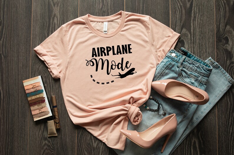 Puede incluir: Una camiseta rosa claro con el texto "AIRPLANE Mode" en letras negras y un peque&ntilde;o gr&aacute;fico de avi&oacute;n. La camiseta est&aacute; colocada sobre una superficie de madera con un par de tacones altos rosa claro y unos vaqueros azules.