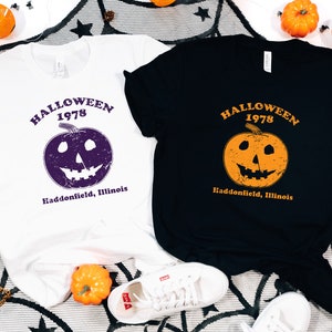 May include: Two t-shirts with a Halloween 1978 design. One shirt is white with a purple pumpkin and the other is black with an orange pumpkin. Both shirts have the text "Halloween 1978" and "Haddonfield, Illinois".