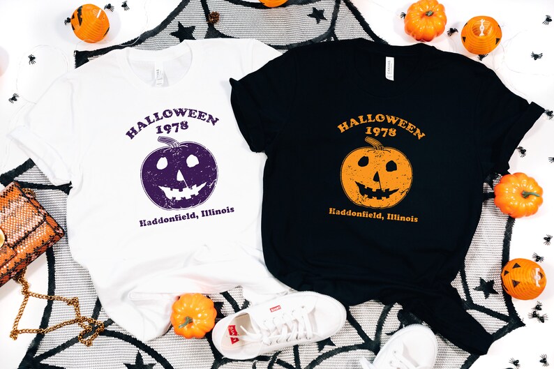 May include: Two t-shirts with a Halloween 1978 design. One shirt is white with a purple pumpkin and the other is black with an orange pumpkin. Both shirts have the text "Halloween 1978" and "Haddonfield, Illinois".