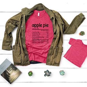 Puede incluir: Una camiseta roja con un gráfico de etiqueta nutricional que dice "apple pie" con la información nutricional enumerada debajo. La camiseta está colocada sobre una superficie de madera blanca con una chaqueta verde y una camiseta roja más pequeña.