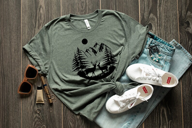 Puede incluir: Una camiseta verde con un dise&ntilde;o gr&aacute;fico negro de monta&ntilde;as, &aacute;rboles y ciervos. Se combina con vaqueros azules y zapatillas blancas. Tambi&eacute;n se ven gafas de sol y accesorios, sobre una superficie de madera.