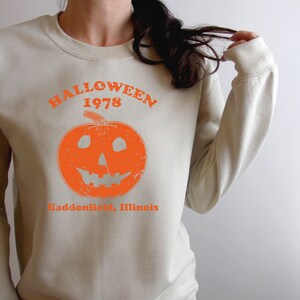 May include: A beige sweatshirt with an orange graphic of a jack-o'-lantern and the text "Halloween 1978 Haddonfield, Illinois".