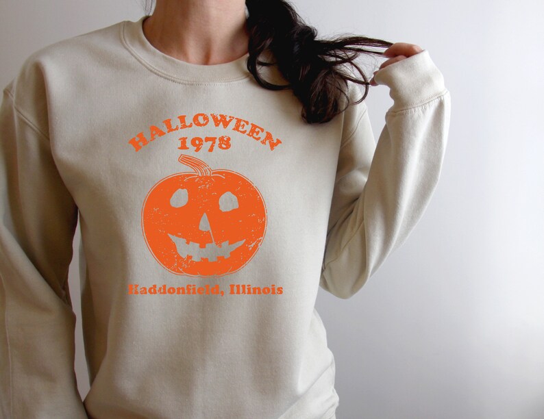 May include: A beige sweatshirt with an orange graphic of a jack-o'-lantern and the text "Halloween 1978 Haddonfield, Illinois".