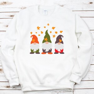 May include: White sweatshirt with a design of three gnomes wearing hats and holding leaves. The gnomes are standing in a row and there are orange leaves falling around them.