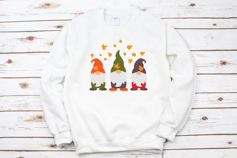May include: White sweatshirt with a design of three gnomes wearing hats and holding leaves. The gnomes are standing in a row and there are orange leaves falling around them.
