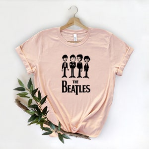Puede incluir: Una camiseta rosa claro con un gr&aacute;fico negro de los Beatles. El gr&aacute;fico presenta cuatro figuras de dibujos animados de los miembros de la banda con trajes, con el texto "THE BEATLES" debajo.