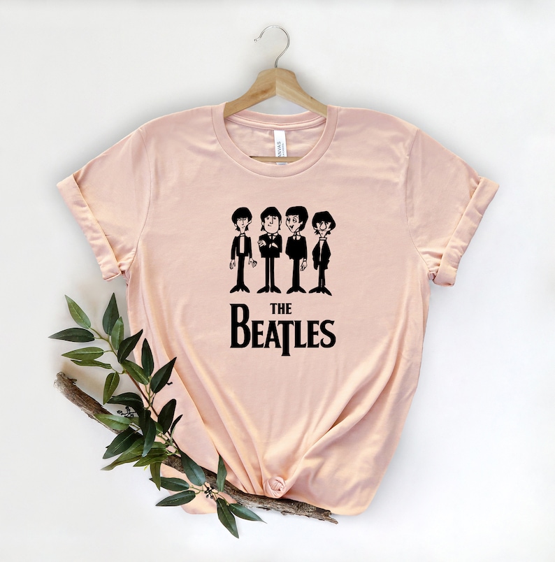Puede incluir: Una camiseta rosa claro con un gr&aacute;fico negro de los Beatles. El gr&aacute;fico presenta cuatro figuras de dibujos animados de los miembros de la banda con trajes, con el texto "THE BEATLES" debajo.