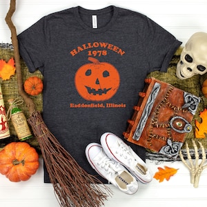 May include: A dark grey t-shirt with a vintage Halloween graphic featuring a jack-o-lantern and the text "Halloween 1978 Haddonfield, Illinois".