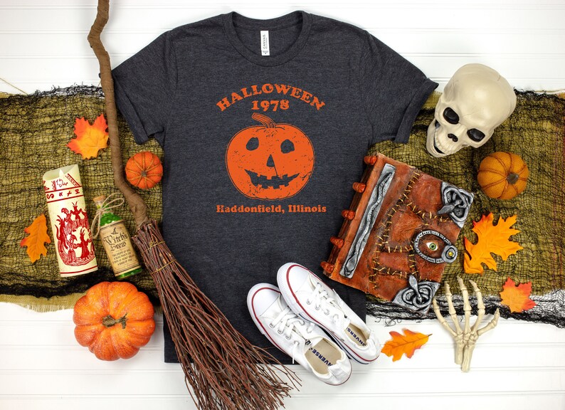May include: A dark grey t-shirt with a vintage Halloween graphic featuring a jack-o-lantern and the text "Halloween 1978 Haddonfield, Illinois".