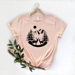 Puede incluir: Camiseta rosa claro con un dise&ntilde;o de silueta negra de dos ciervos, &aacute;rboles, monta&ntilde;as y un sol. La camiseta se muestra en una percha de madera. Las mangas est&aacute;n enrolladas.
