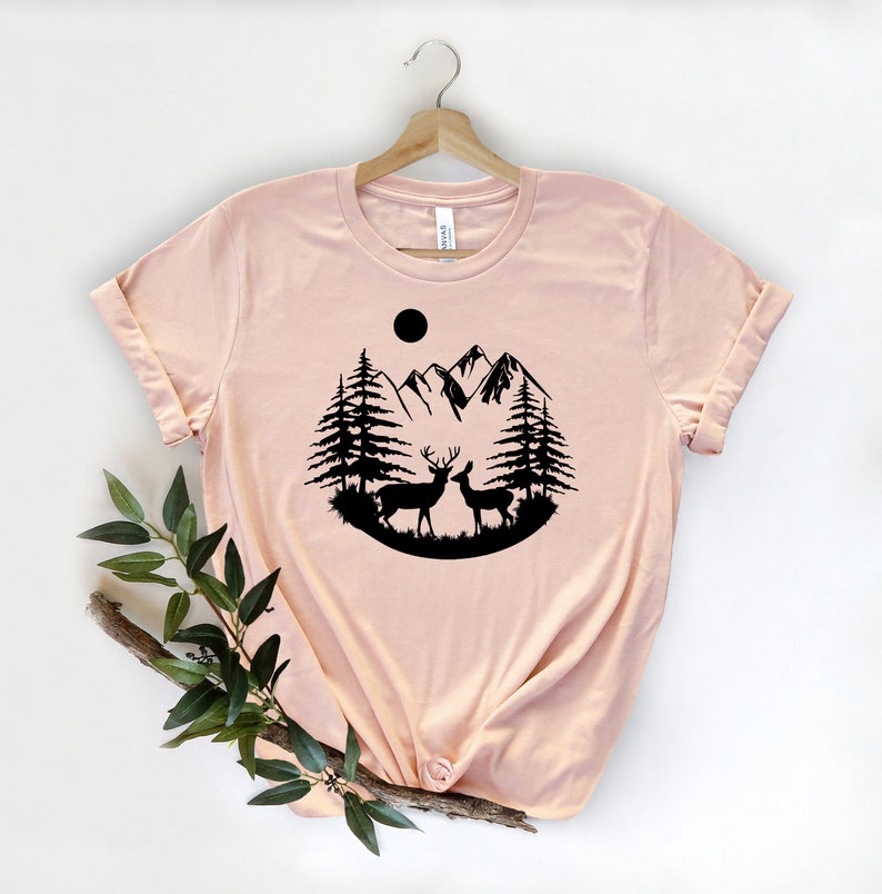 Puede incluir: Camiseta rosa claro con un dise&ntilde;o de silueta negra de dos ciervos, &aacute;rboles, monta&ntilde;as y un sol. La camiseta se muestra en una percha de madera. Las mangas est&aacute;n enrolladas.