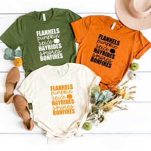 May include: Three t-shirts with the text "FLANNELS pumpkin spice HAYRIDES smores BONFIRES" printed on them. The shirts are in green, orange, and cream colours.