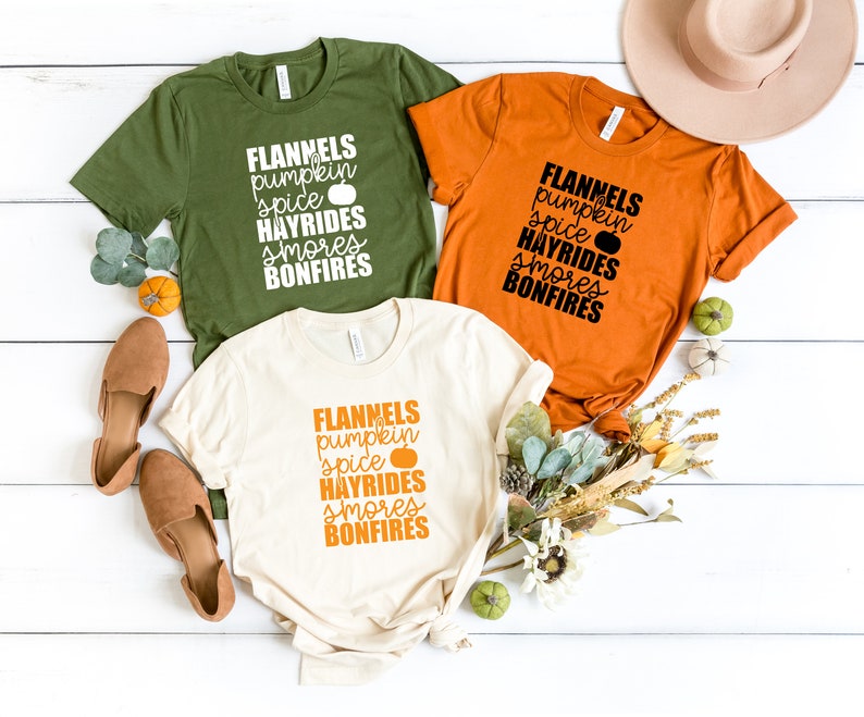 May include: Three t-shirts with the text "FLANNELS pumpkin spice HAYRIDES smores BONFIRES" printed on them. The shirts are in green, orange, and cream colours.