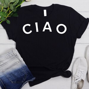 May include: Black t-shirt with white text that says "I CIAO". The shirt is laying on a white wooden surface with a pair of blue denim shorts and white sneakers.