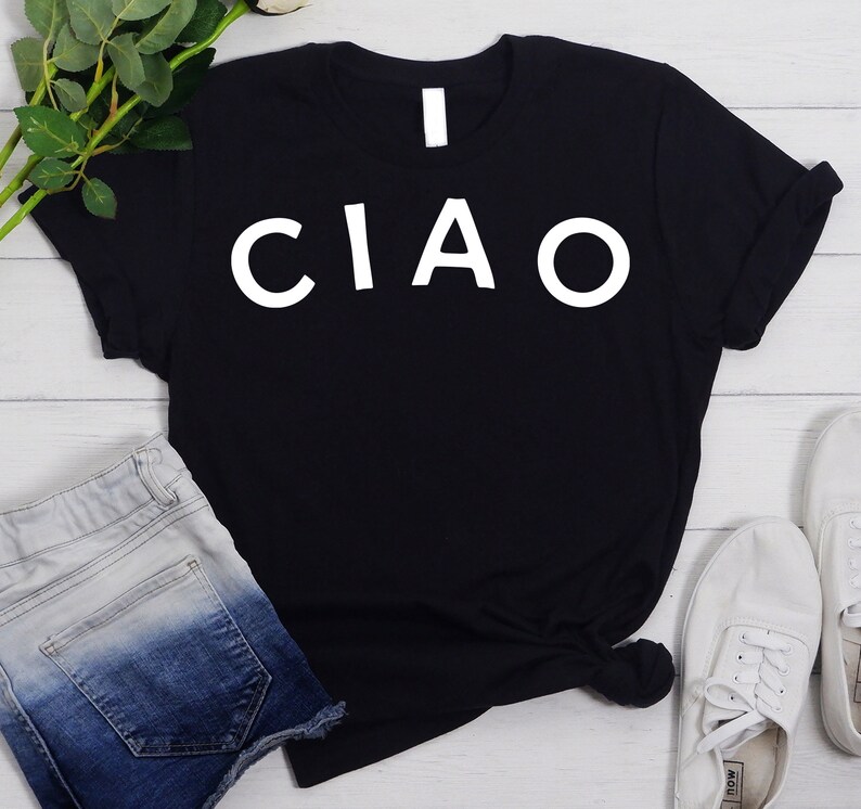 May include: Black t-shirt with white text that says "I CIAO". The shirt is laying on a white wooden surface with a pair of blue denim shorts and white sneakers.
