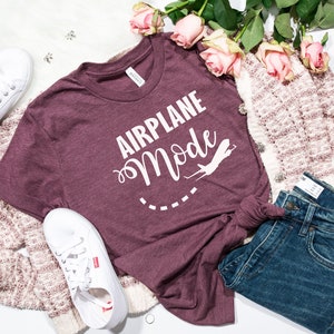 Puede incluir: Una camiseta de color granate con el texto "Airplane Mode" en letras blancas y un gr&aacute;fico de avi&oacute;n blanco. La camiseta est&aacute; colocada sobre una superficie blanca con un par de zapatillas blancas, un par de vaqueros azules y una chaqueta de punto crema y rosa.