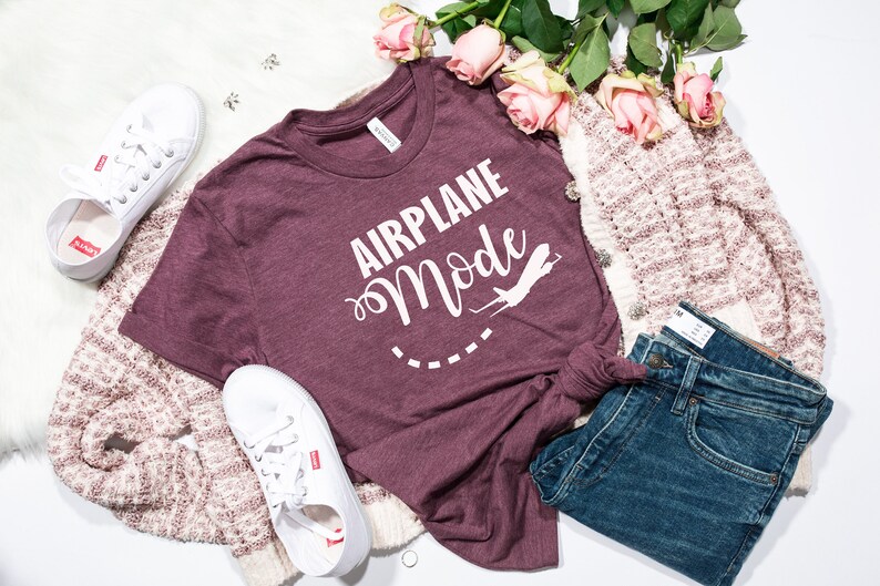 Puede incluir: Una camiseta de color granate con el texto "Airplane Mode" en letras blancas y un gr&aacute;fico de avi&oacute;n blanco. La camiseta est&aacute; colocada sobre una superficie blanca con un par de zapatillas blancas, un par de vaqueros azules y una chaqueta de punto crema y rosa.