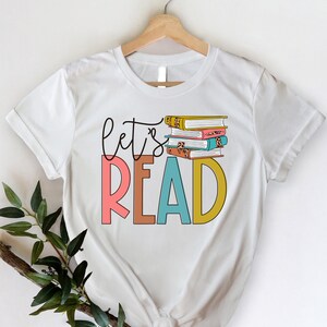 May include: White t-shirt with the text "let's read" in colorful letters and a stack of books with colorful covers.