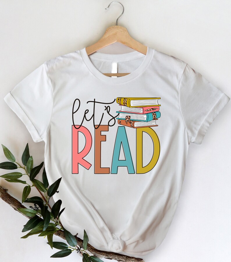 May include: White t-shirt with the text "let's read" in colorful letters and a stack of books with colorful covers.