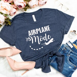 Puede incluir: Una camiseta azul marino con el texto "Airplane Mode" en letras blancas. Un peque&ntilde;o gr&aacute;fico de avi&oacute;n est&aacute; al lado del texto. La camiseta est&aacute; colocada sobre una superficie blanca con un par de jeans, un par de tacones altos y una botella de perfume.