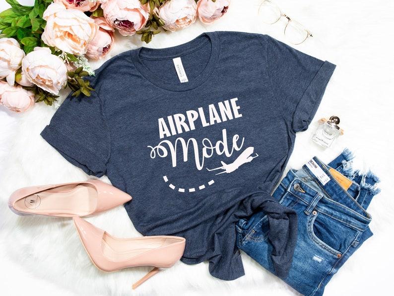 Puede incluir: Una camiseta azul marino con el texto "Airplane Mode" en letras blancas. Un peque&ntilde;o gr&aacute;fico de avi&oacute;n est&aacute; al lado del texto. La camiseta est&aacute; colocada sobre una superficie blanca con un par de jeans, un par de tacones altos y una botella de perfume.