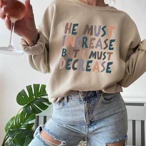 K&ouml;nnte beinhalten: Ein beiger Pullover mit dem Aufdruck "HE MUST INCREASE BUT I MUST DECREASE" in bunten Buchstaben. Der Text ist in einem verspielten, handgeschriebenen Stil.