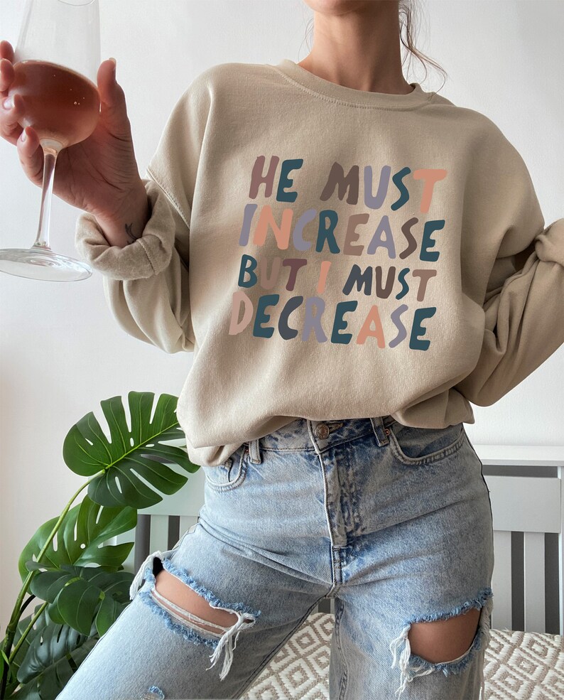 K&ouml;nnte beinhalten: Ein beiger Pullover mit dem Aufdruck "HE MUST INCREASE BUT I MUST DECREASE" in bunten Buchstaben. Der Text ist in einem verspielten, handgeschriebenen Stil.