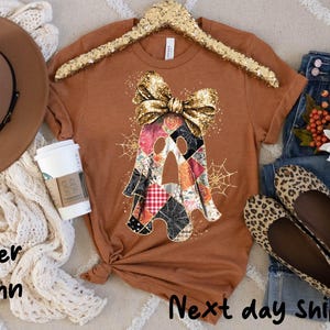 May include: A burnt orange t-shirt with a patchwork ghost design, a gold sequined bow, and spiderwebs. The shirt is displayed with a brown fedora, a cream-colored scarf, jeans, and leopard print flats. A coffee cup is also present.