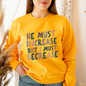 K&ouml;nnte beinhalten: Gelber Pullover mit dem Text "He must increase but I must decrease" in bunten Buchstaben.