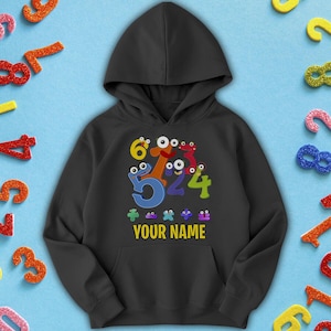May include: Black hooded sweatshirt with a playful design of cartoon numbers with eyes. The numbers 6, 5, 2, 4, and 3 are featured, along with the text "YOUR NAME" in a yellow box. The sweatshirt has a front pocket and a hood.