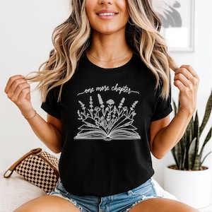 May include: A black short-sleeved t-shirt featuring a white line art graphic of an open book with various wildflowers growing out of the pages. Above the book, the text reads one more chapter in a cursive script font. The shirt has a crew neckline.