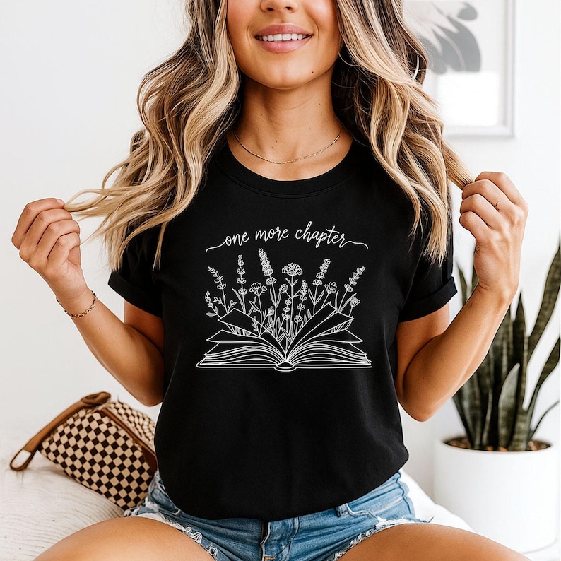 May include: A black short-sleeved t-shirt featuring a white line art graphic of an open book with various wildflowers growing out of the pages. Above the book, the text reads one more chapter in a cursive script font. The shirt has a crew neckline.