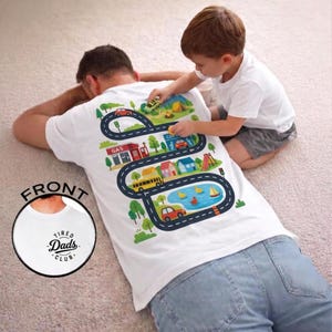 May include: A white t-shirt featuring a back graphic of a winding road with cars, a petrol station, houses, and a pond. The front chest area displays a circular logo with the text Tired Dads Club. The design includes green trees and colourful buildings.