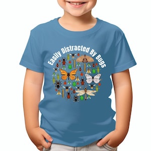 May include: A toddler wearing a light blue t-shirt with the text "Easily Distracted By Bugs" in white. The text is surrounded by a circle of colourful bugs, including butterflies, beetles and bees.