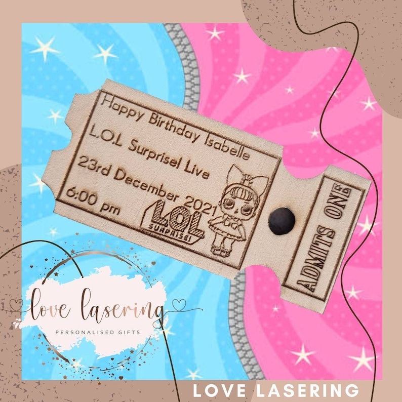 May include: A wooden ticket with a brown background and white text. The ticket says "Happy Birthday Isabelle LOL Surprise! Live 23rd December 2021 6:00 pm" and "ADMITS ONE". The ticket has a small image of a LOL Surprise! doll on it.